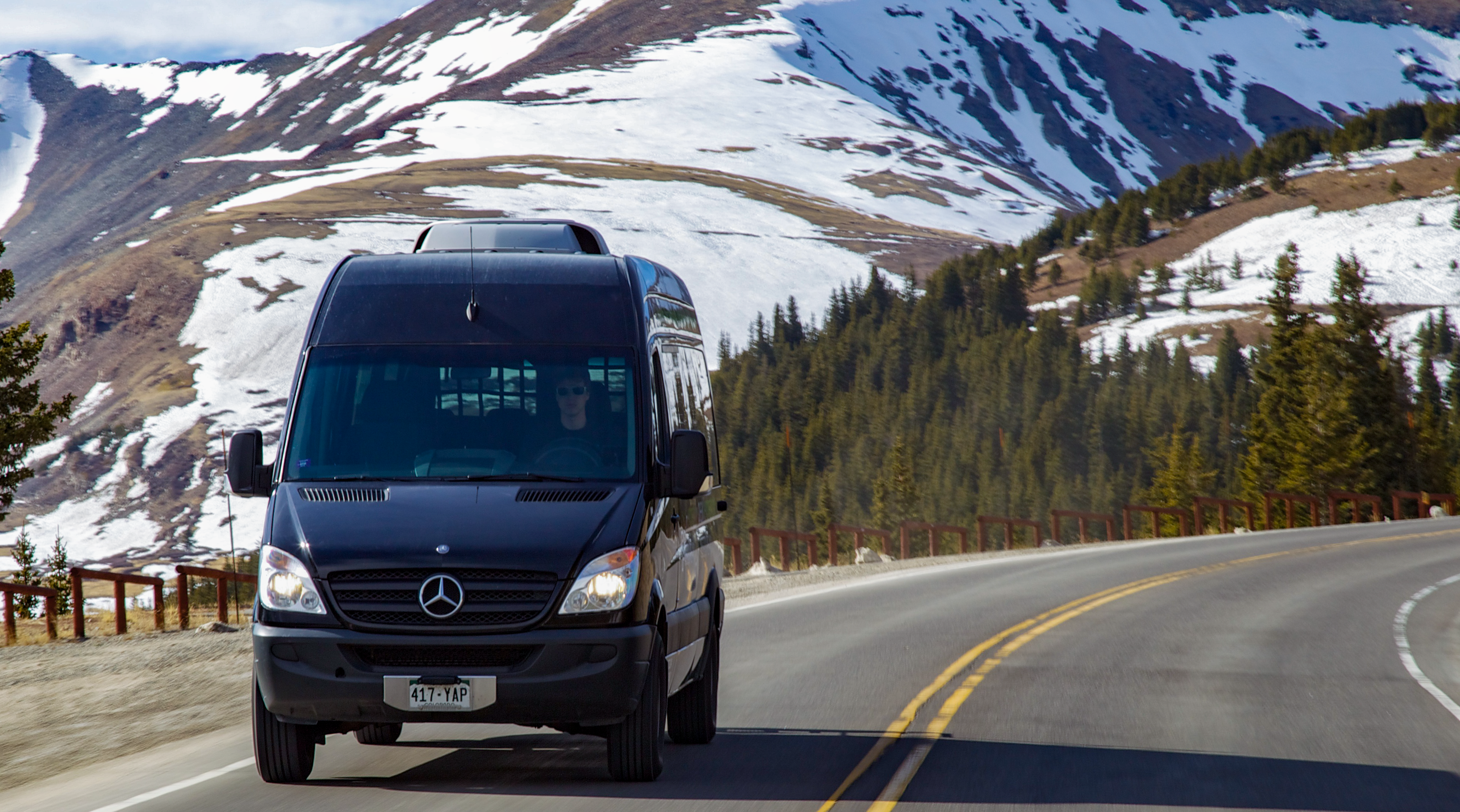 A black sprinter van driving on a road with mountains in the background