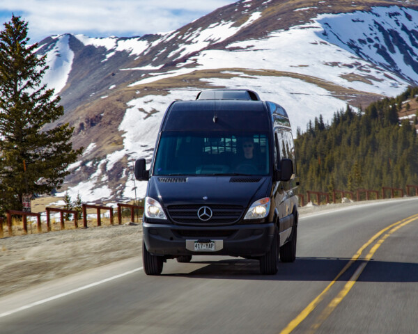 A black sprinter van driving on a road with mountains in the background