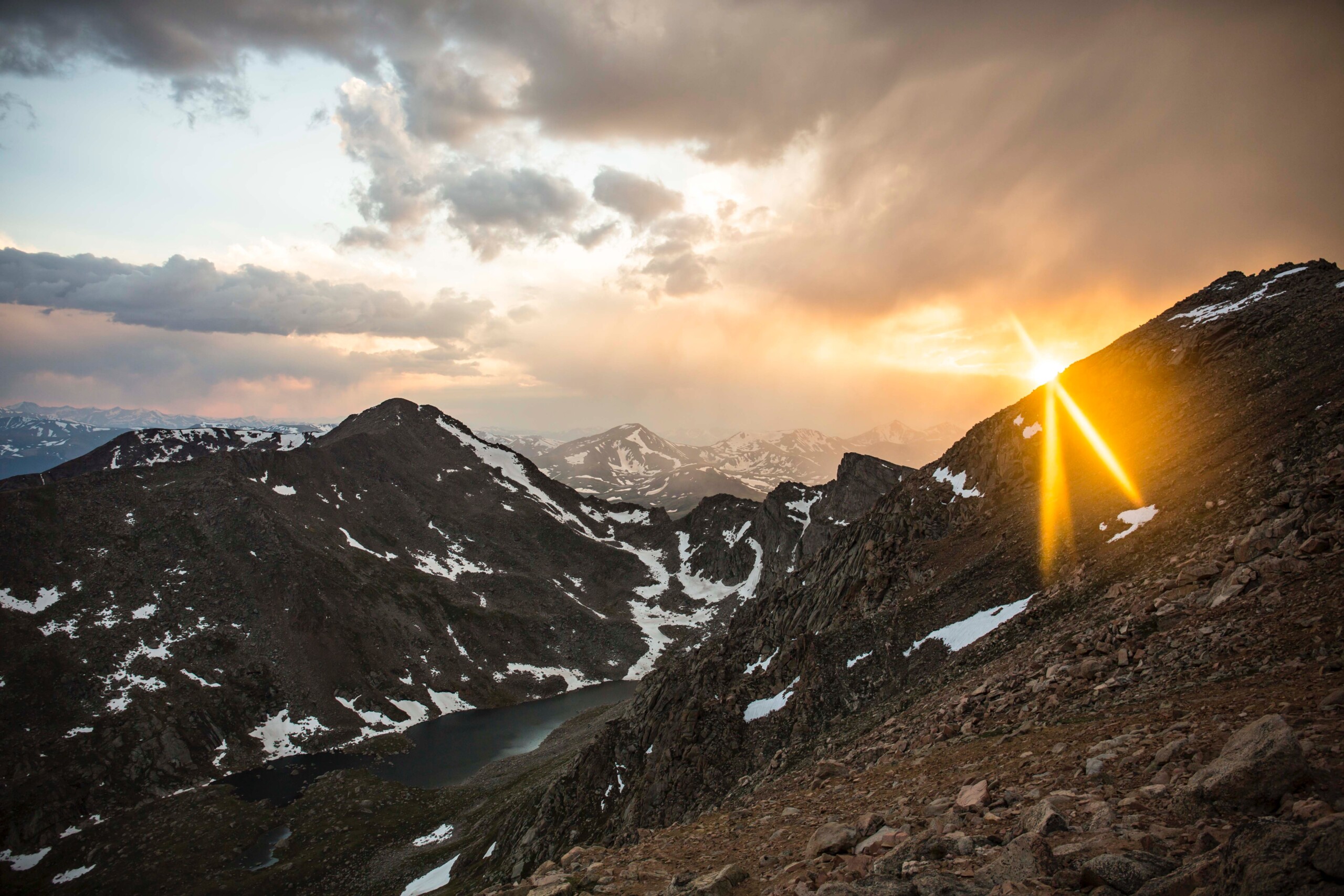 The peaks of mountains and a lake nestled among them with the sun glaring from just above the ridgeline